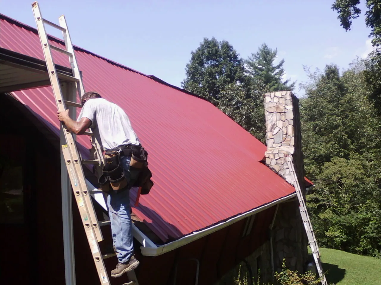 Roofer on a ladder beside a red metal roof and stone chimney — Soffit & Fascia Repair by Roofing Brothers in Glencliff